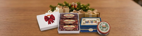 White fruitcake gift box and holiday gift tins arranged beside evergreen greenery and warm Christmas lights