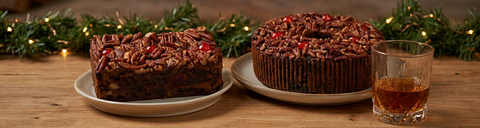 Bourbon fruitcake loaf and ring topped with pecans and cherries, beside a low glass of bourbon on a rustic wooden table.