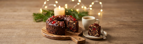 Dark fruitcake topped with pecans and cherries, with a plated slice and coffee beside holiday greenery and candlelight