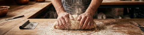 Baker kneading dough on a flour-dusted wooden worktop, with airborne flour illuminated by warm side lighting.