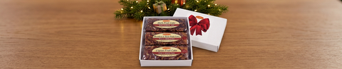 White gift box containing three individually wrapped 1-pound fruitcakes, with a red-bow lid beside holiday greenery and lights.