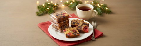 Fruitcake slices on a white plate with individually wrapped portions, tea, and a dessert fork beside holiday greenery and lights.