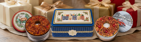 Blue winter-village gift tin between two open fruitcake tins with deer and red-truck lids, surrounded by wrapped presents.
