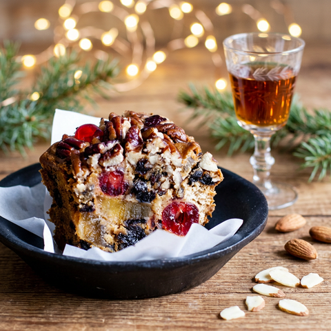 Amaretto fruitcake slice on a dark plate with whole and sliced almonds, a glass of amaretto, evergreen sprigs, and warm holiday lights