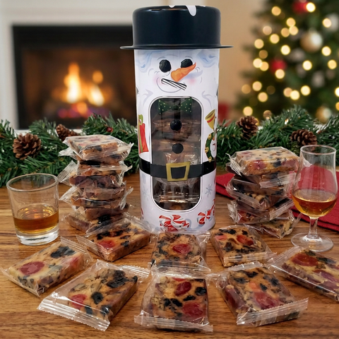 Individually wrapped Jane Parker Bourbon & Rum fruitcake slices around a tall snowman tin, beside two glasses of amber spirits in a Christmas setting