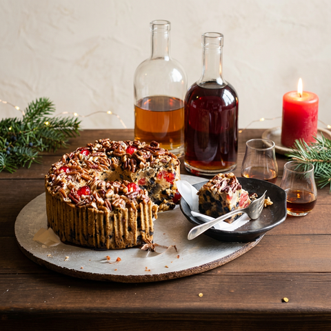 Bourbon & Rum ring fruitcake with a slice on a dark plate, bourbon and rum bottles, small tasting glasses, evergreen sprigs, and a glowing red candle