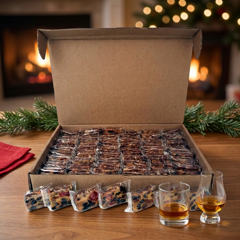 Open bulk box of individually wrapped Jane Parker Bourbon & Rum fruitcake slices beside two glasses of amber spirits on a wooden table with Christmas greenery