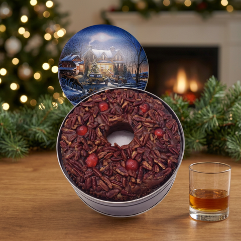 Jane Parker Bourbon Fruitcake ring in an open holiday tin with a snowy-house lid, beside a small glass of bourbon and holiday greenery