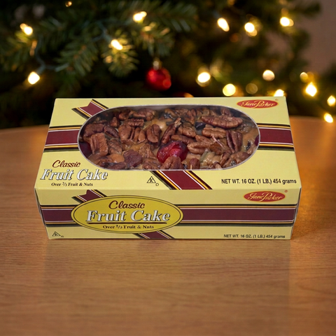 Jane Parker Classic Fruit Cake 1 lb. yellow window box on a wooden table, with a softly blurred Christmas tree and warm white lights behind.