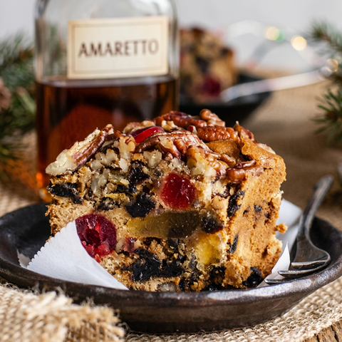 Close-up of Amaretto fruitcake with cherries, raisins, pineapple, and pecans on a plate, with a partially filled amaretto bottle behind it