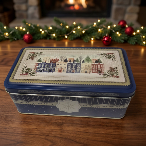 Closed blue holiday fruitcake tin with winter village artwork on a wooden table, with evergreen garland, red ornaments and warm lights
