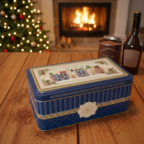 Closed blue holiday tin with winter village artwork on a cocoa-dusted wooden table, with a cocoa tin, liqueur bottle, fireplace and lit Christmas tree behind
