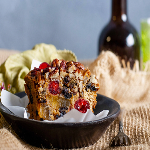 Bourbon & Rum fruitcake slice with cherries, raisins, pineapple, and pecans in a parchment-lined dark bowl, with a bottle in the background