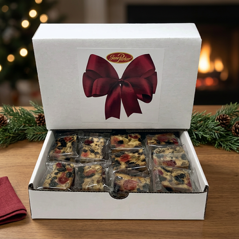 Open Jane Parker gift box of individually wrapped Classic fruitcake slices, with a burgundy bow design, on a wooden table with Christmas greenery and a fireplace.