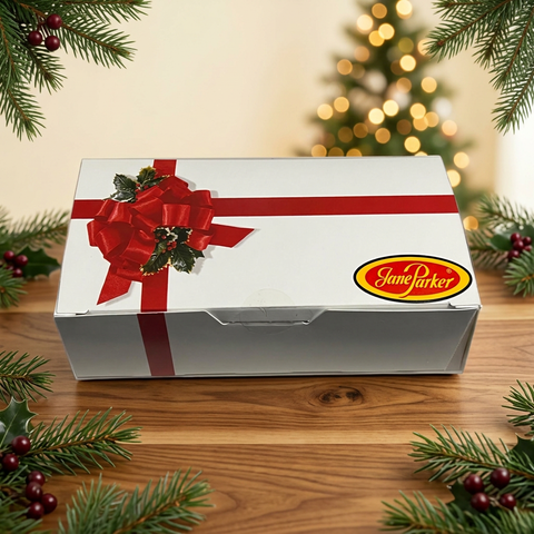 Jane Parker white holiday gift box with a red ribbon design on a wooden table, surrounded by evergreen branches and softly glowing Christmas lights