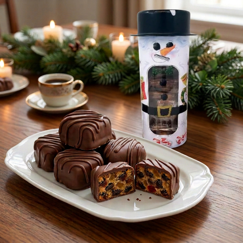 Milk chocolate-covered fruitcake slices on a white platter beside a snowman gift tin, with coffee and festive greenery