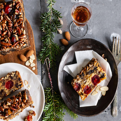 Overhead view of Amaretto fruitcake and slices with whole and sliced almonds, a glass of amaretto, and holiday greenery