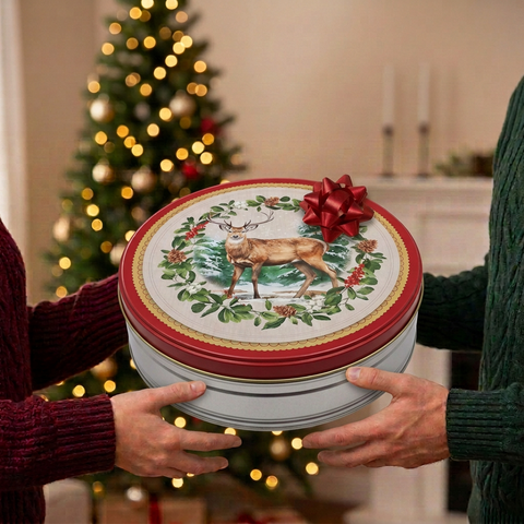Two people exchanging a shallow round holiday fruitcake tin with deer artwork and a small red bow beside a softly lit Christmas tree