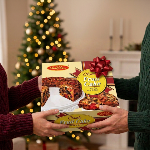 Two people in burgundy and green sweaters exchanging a Jane Parker Classic Fruit Cake box with a red bow beside a softly lit Christmas tree