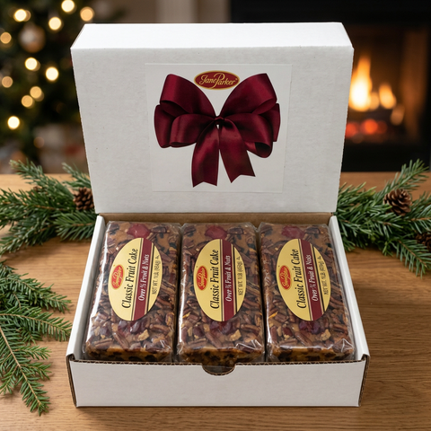 Three individually wrapped Classic Fruit Cake 1 lb. loaves aligned in an open white gift box with a burgundy bow design, beside greenery and a glowing fireplace