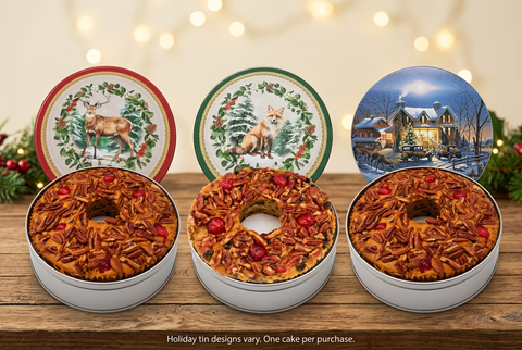 Amaretto ring fruitcakes displayed with deer, fox, and snowy-house holiday tin lids. Tin designs vary; one cake per purchase
