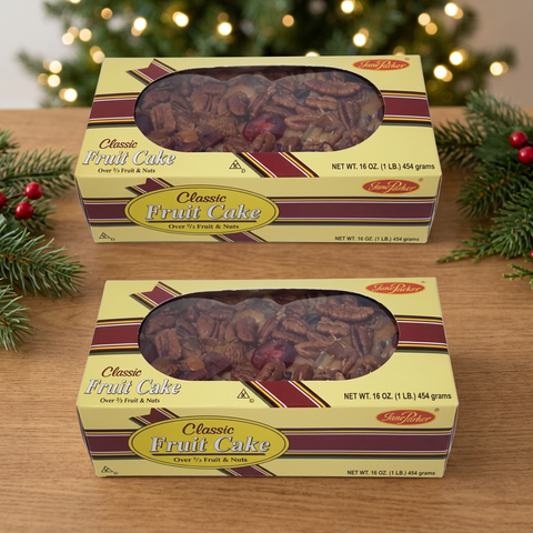 Two Jane Parker Classic Fruit Cake 1 lb. yellow window boxes on a wooden table, framed by evergreen sprigs and softly glowing Christmas lights.