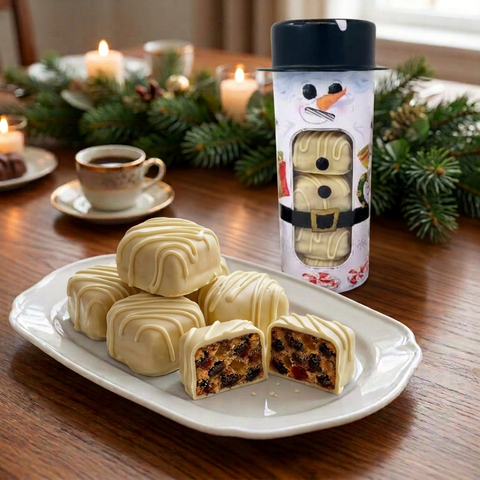 White chocolate-covered fruitcake slices on a white platter beside a snowman gift tin, with coffee and festive greenery